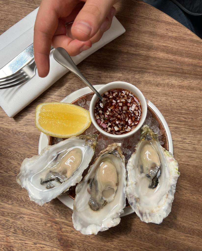 Plate of fresh oysters on ice with a wedge of lemon and a small bowl of sauce, accompanied by a hand holding a spoon.