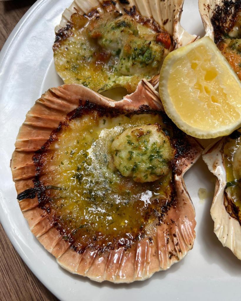 Grilled scallops in their shells, topped with herbs and breadcrumbs, served with a lemon wedge.