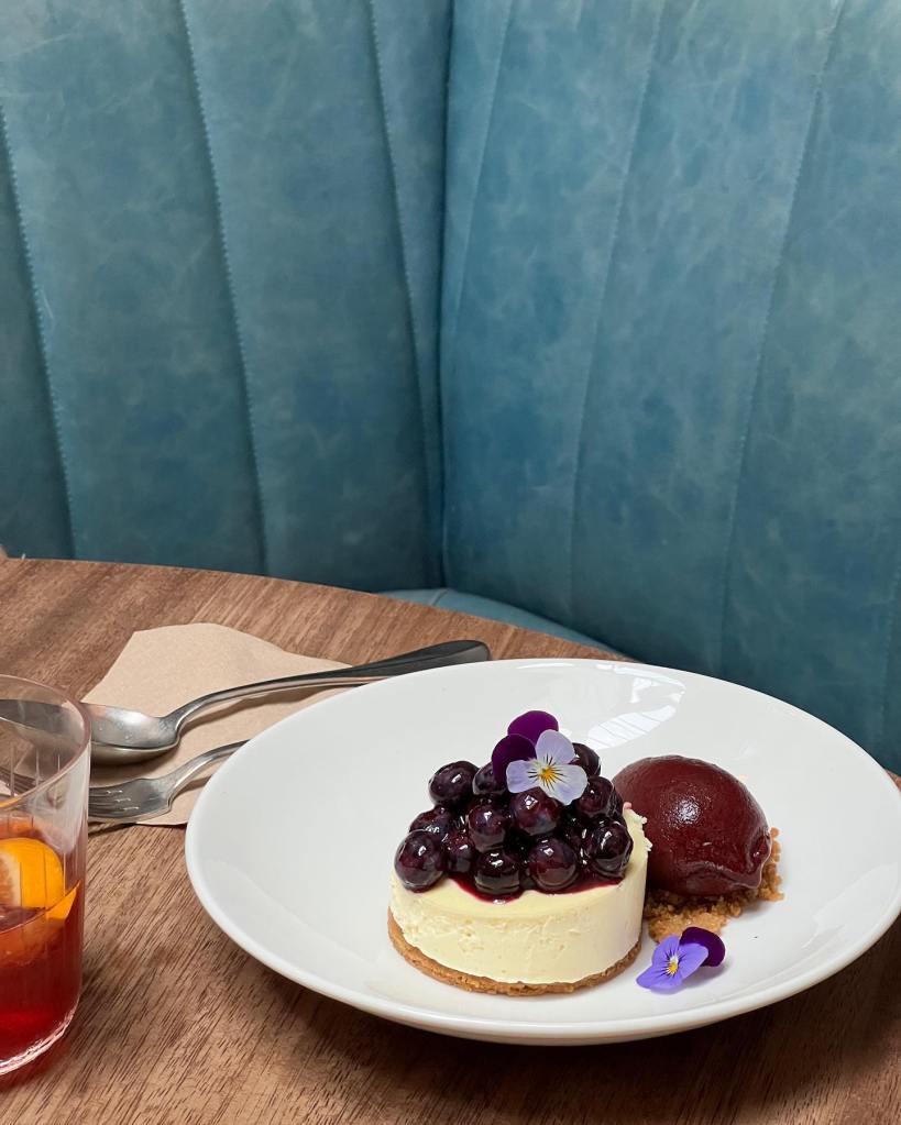 A plate featuring a cheesecake topped with blueberries and edible flowers, alongside a scoop of berry sorbet, with a glass of beverage in the background on a wooden table.