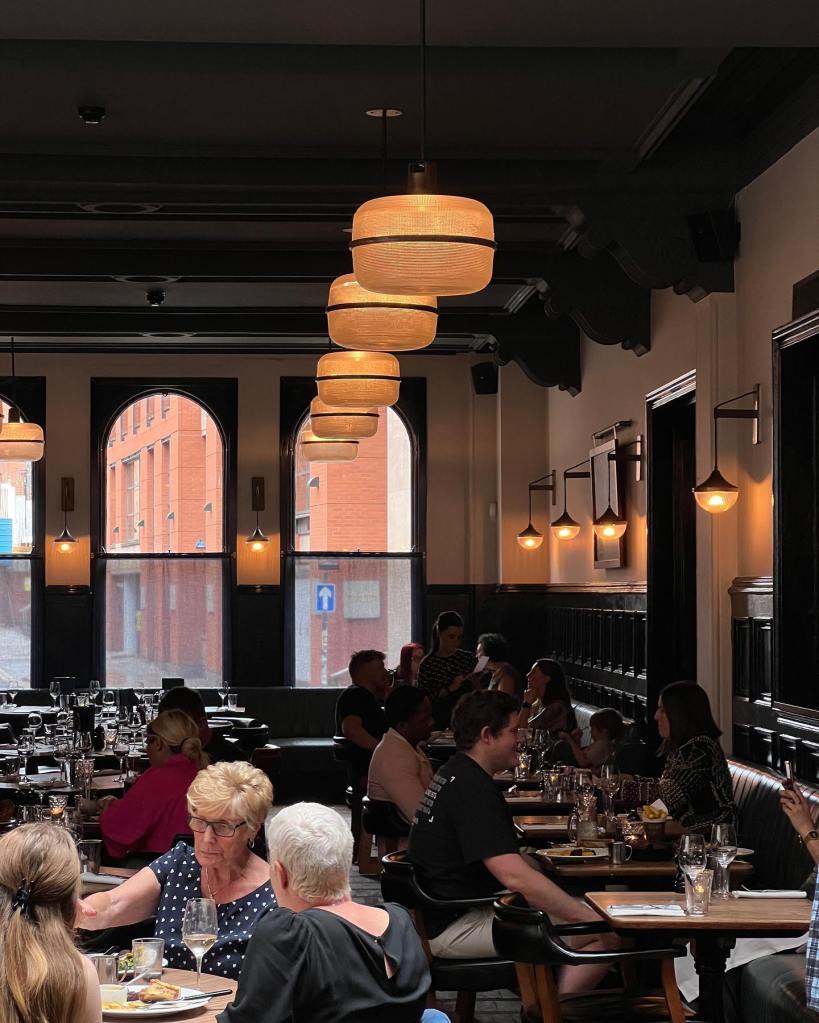 A lively restaurant interior featuring people dining at tables, elegant pendant lights, and large windows. The decor blends modern and traditional elements, with a warm ambiance.