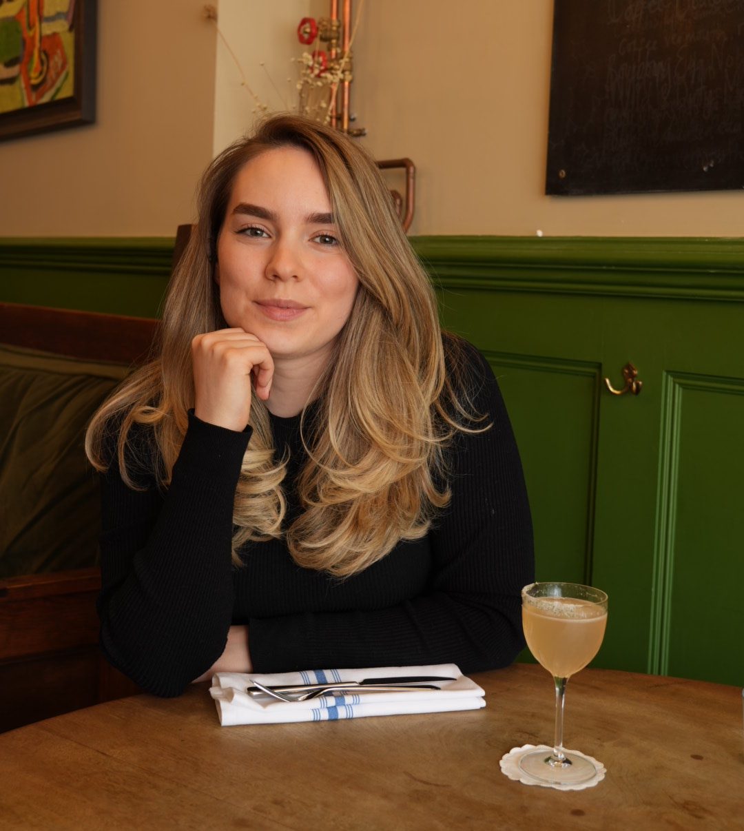 A young woman with long, wavy hair sits at a wooden table, resting her chin on her hand. She smiles softly while looking directly at the camera. A cocktail is placed in front of her along with a neatly arranged fork and knife on a napkin.