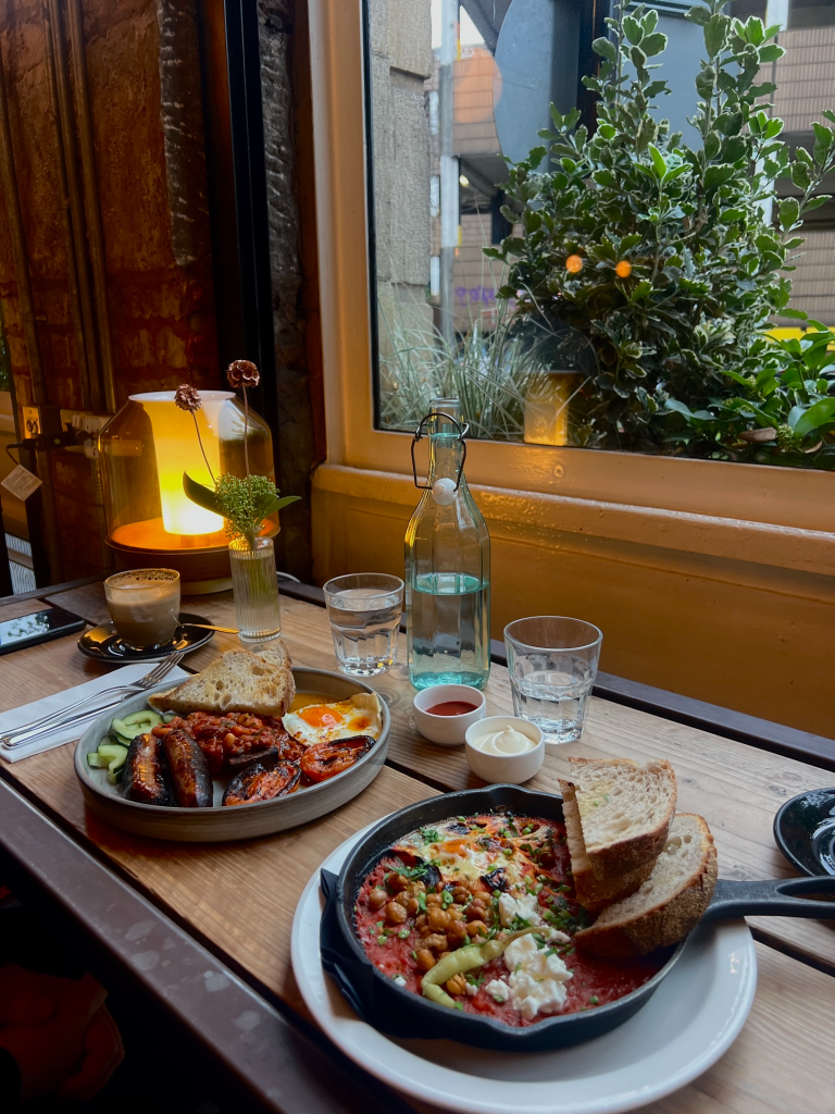 A cozy breakfast table featuring a skillet of baked beans with eggs, and a side plate with grilled sausages, cucumbers, and toast. A glass bottle of water, two glasses, and a cup of coffee are also on the wooden table, with greenery visible outside the window.