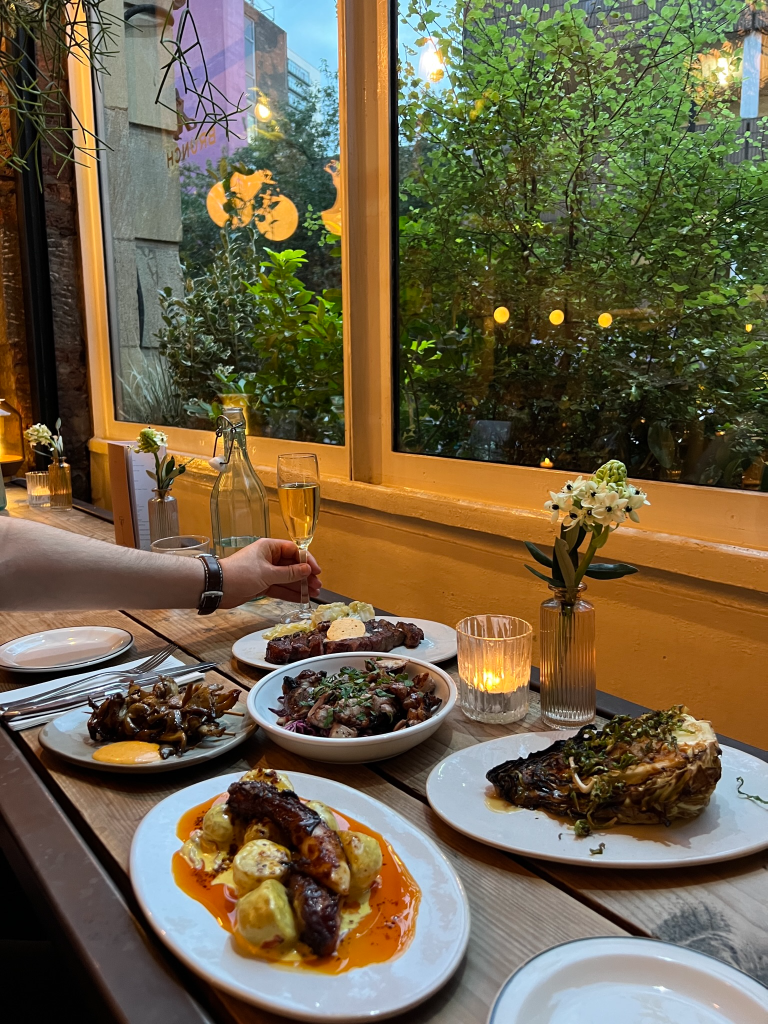 A dining table set with several plates of gourmet food, including grilled meats, roasted vegetables, and garnishes, with a hand holding a glass of champagne. The setting features a large window with greenery outside and soft lighting.