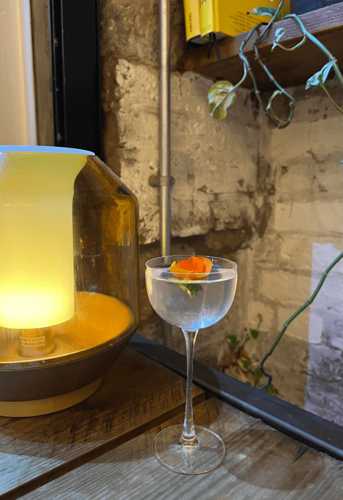 A cocktail glass filled with a clear drink and garnished with a small orange flower, placed next to a warm, glowing lamp on a wooden table.