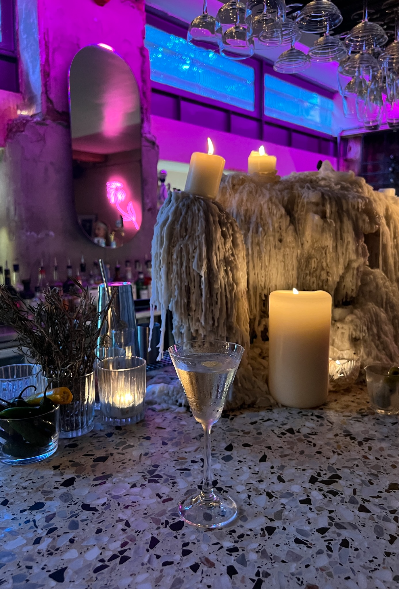 A dimly lit bar scene featuring a cocktail glass with a lemon twist, surrounded by candles and decorative elements, with pink and blue lighting.