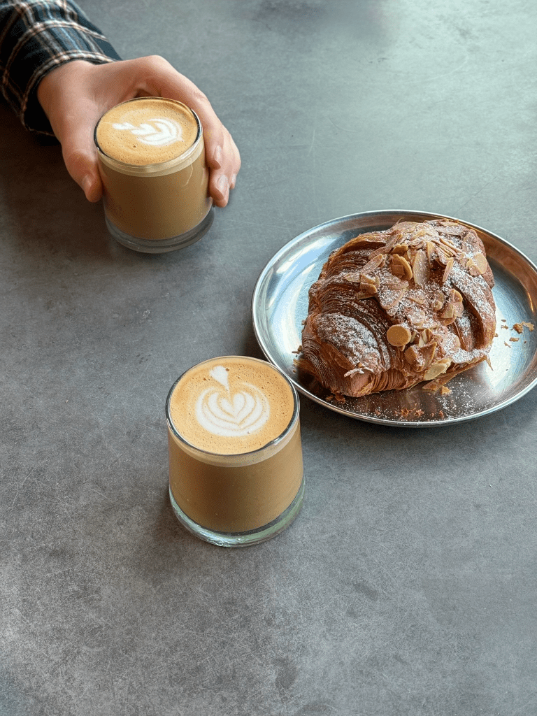 A hand holding a glass of latte with latte art, next to a plate of almond croissant on a gray table.